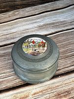 Closed view of round metal music box with hunting scene on lid