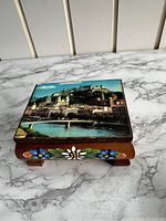 Top view of vintage wooden music trinket box showing scenic image of a castle and town on the lid and hand-painted flowers on the side.