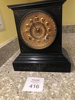 Front view of clock on countertop showing black wooden case, brass dial, and gold inlay scrollwork.