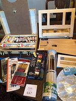 Overview showing wooden lap easel, paint tubes laid in a wooden box organizer, several packs of paint brushes, a round palette, and a cylindrical container holding 24 brushes.