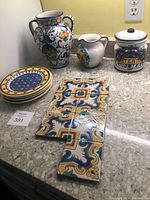 Image showing ceramic urn, pitcher, tea container, plates, and decorative tiles arranged on countertop.