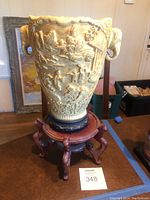 Full view of ivory resin vase on wood stand including relief carvings and elephant handles