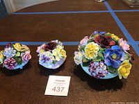 Overall view of three assorted hand-modeled bone china floral bouquets in various sizes and colors arranged on a brown surface.