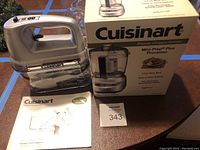Cuisinart 9-speed hand mixer HM-90S with storage case and Cuisinart Premier chopper/grinder in box, placed on wooden table.