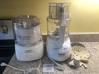 Front view of white food processor and frozen yogurt maker with cords and accompanying accessories including slicing/shredding disc and dough blade on kitchen counter.