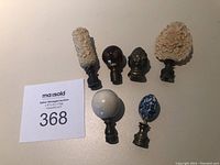 Top view of six finials, showing varied shapes and materials including cream textured, dark bulb, metal face, large floral, silver sphere, and blue-white ceramic.