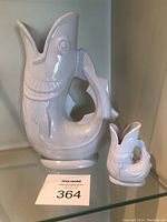 Wide shot showing both large and small white ceramic gurgling cod pitchers on a glass shelf.