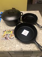 Set of three cast iron items including a Dutch oven with lid and two frying pans on a kitchen counter along with a Lodge Cast Iron booklet.