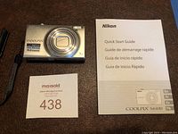 Nikon Coolpix S6100 silver camera shown with wrist strap and quick start guide manual on brown surface.