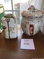 Two ceramic pieces placed on a table: a rectangular vase with pink and purple floral designs and a round cookie jar with lid featuring varied floral patterns and gold detailing.