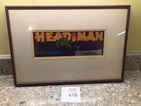 Full view of framed advertisement with beige mat and reddish brown frame showing 'HEADMAN' brand text and rooster graphic on dark blue.