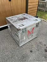Metal square flight/road case with visible wear and a red letter M on the side, photographed outdoors on asphalt near wooden doors.
