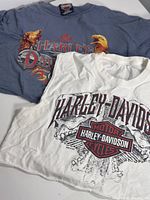 Two women's crop tops, one blue-gray with eagle graphic and another white with skull wings and Harley-Davidson logo.