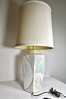 Front side view of the large table lamp showing the opal iridescent ceramic base and fabric shade, lamp plugged in with cord coiled beside base.