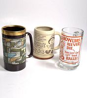 Photo showing three vintage beer steins side by side on a white background. Left is pottery stein with abstract geometric design and gold rim, middle is light brown ceramic stein with humorous cartoons and text, right is clear glass stein with red bowling humor.