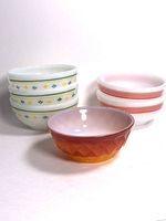 Six vintage milk glass bowls arranged showing Fire King amber-orange bowl front and center with five white Termicrisa bowls behind it with floral and red ring decorations