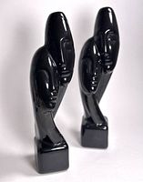 Two black glossy Art Deco sculptures with elongated stylized human faces shown upright with square bases.