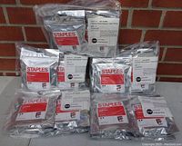 Photo showing 9 sealed silver foil packages of Canon PGI-5 black ink cartridges from Staples labeled SIC-R50B2, arranged on a brick floor, highlighting the packaging and quantity.