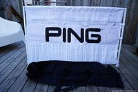 Front view of white Ping branded golf club display stand with 20 slots for golf clubs and detachable black carry bag below.
