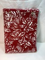 Image of folded tablecloth in original packaging showing red fabric with white holiday-themed botanical print including poinsettias, pine cones, and berries.