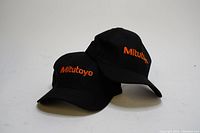 Pair of two black baseball hats with orange Mitutoyo logo, shown stacked.