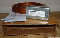 Rectangular metal housing labeled Siemens QAM2012.750 with coiled copper wire probe and instruction papers on brown cardboard box.