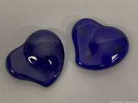 Two cobalt blue glass heart-shaped paperweights placed side-by-side with reflections visible on their surfaces, showing shape and color.