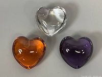 Top view of three heart-shaped paperweights: clear, amber, and purple, showing shape and color.