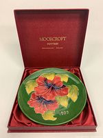 Top-down view of ceramic plate with red and pink hibiscus flowers, green background, inside red Moorcroft box with satin lining, year 1983 on plate
