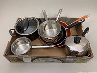 Box of assorted pots and pans showing metal and non-stick materials with various handles and lids