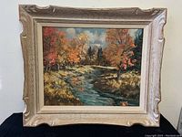 Frontal view of the framed painting showing the river landscape with autumn trees and the ornate frame with minor wear.