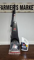 Full upright view of Bissell Quicksteamer Powerbrush multi-surface cleaner with attached cord and nozzle, next to jug of Hoover Oxy Permanent Stain Remover.