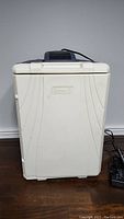 Closed white Coleman electric cooler with vented black panel on top and power cord coiled beside it.