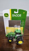 Front view of John Deere 8400R die-cast tractor toy in packaging showing the tractor inside a clear plastic blister and the branded card backing with model number and scale details.