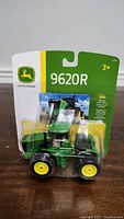 Front view of the John Deere 9620R 1/64 die-cast metal model in blister packaging showing detailed tractor body and wheels.
