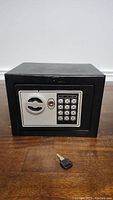 Front view of the small black safe box with keypad and key placed on wooden surface.