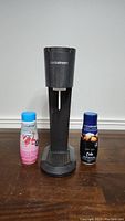 Front view of black Sodastream soda maker machine with Sodastream logo, alongside two flavor syrup bottles, strawberry kiwi and dry ginger cola.