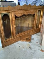 Front view of the wooden and glass corner religious icon cabinet showing carved wood details, glass door, cross designs on side glass panels, and two small drawers at base.