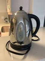 Photo of stainless steel electric kettle showing reflective body, black handle, water level gauge, and base with power cord.
