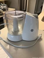 Side view of GE food processor showing white base, clear bowl with blade, power cord, and GE logo