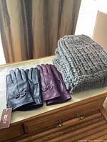 Two pairs of leather gloves, black and burgundy, new with tags, next to a grey hand knit hat and matching scarf set on a tabletop.