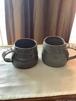 Two matching grey ceramic coffee mugs shown side by side on a light surface with curtain background, showing the round shape and handles, no chips or scratches visible.
