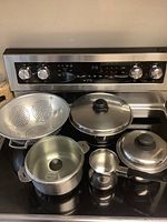 Photo showing all items on stovetop: three stainless steel pots/pans with lids, aluminum colander, and Turkish coffee pot.