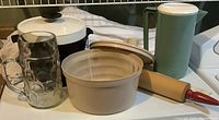 Photo shows five kitchen items: a beige plastic salad spinner with white lid, a translucent dark brown ice bucket with white lid, a wooden rolling pin with red handles, a green plastic juice container with white lid, and a large glass beer stein with handle and dimpled surface