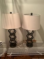 Pair of matching table lamps with black sculptural metal bases and white drum shades on a white wire shelf.