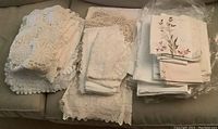 Top-down view of various small folded table cloths and doilies in white and cream tones, with one stack showing floral embroidery, on a grey couch