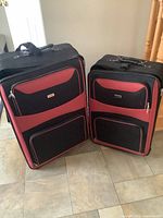 Two black and red patterned softside luggage pieces standing upright on a tiled floor near stairs, showing overall size difference and front zipper pockets.