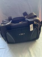 Front view of brand new black overnight bag with visible tag attached and logo plate on front pocket.