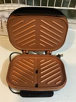 Opened view showing the copper-colored non-stick ridged cooking plates, square surface approximately 9 by 9 inches, with two drainage holes.
