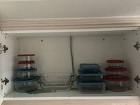 Various sizes of glass Pyrex and Anchor Hocking containers with rubber lids, two rectangular glass bakeware dishes arranged in a cabinet.